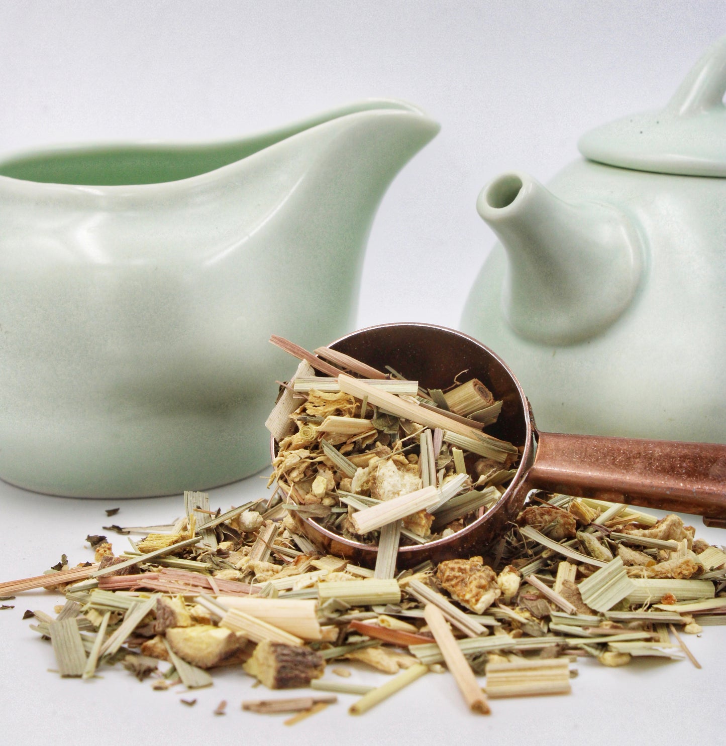Organic Lemon Ginger Tea blend by Relaxation Tea showcased in a copper scoop with loose-leaf herbs spilling onto a clean surface, accompanied by elegant light green teapot and creamer. Featuring organic ginger, lemongrass, lemon peel, licorice root, and spearmint, this zesty and soothing blend is crafted to support digestion and relaxation, highlighting Relaxation Tea’s dedication to organic wellness solutions and premium-quality tea.