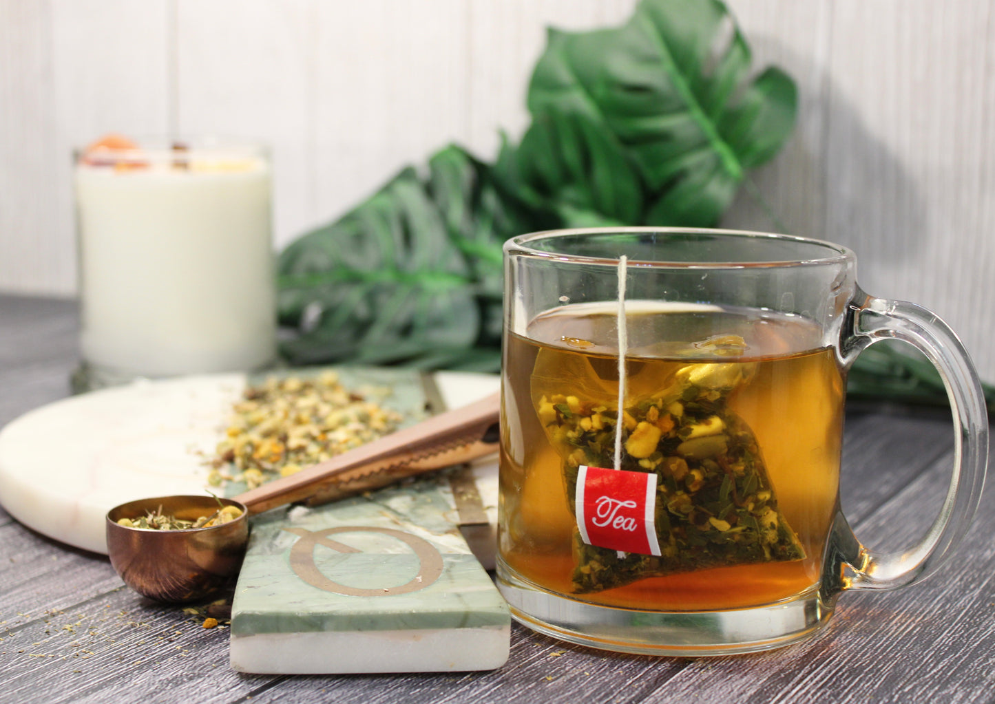 Digestive Tea blend by Relaxation Tea steeping in a glass mug, accompanied by a copper scoop of loose-leaf herbs on a marble board with greenery and a candle in the background. This beautifully styled setup highlights the tea’s natural ingredients and soothing properties, crafted to support healthy digestion and promote gut wellness, showcasing Relaxation Tea’s dedication to high-quality herbal remedies for overall health.