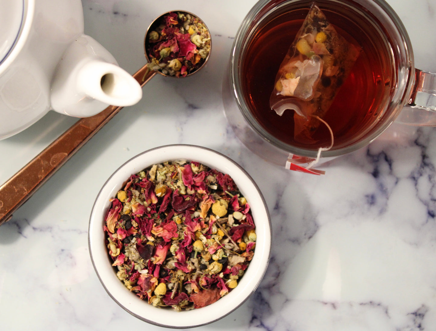 Top-down view of a marble countertop with a glass mug of breath easy respiratory herbal tea, a loose tea strainer with dried flowers and herbs, and a white teapot, showcasing a calming and aesthetic tea preparation.