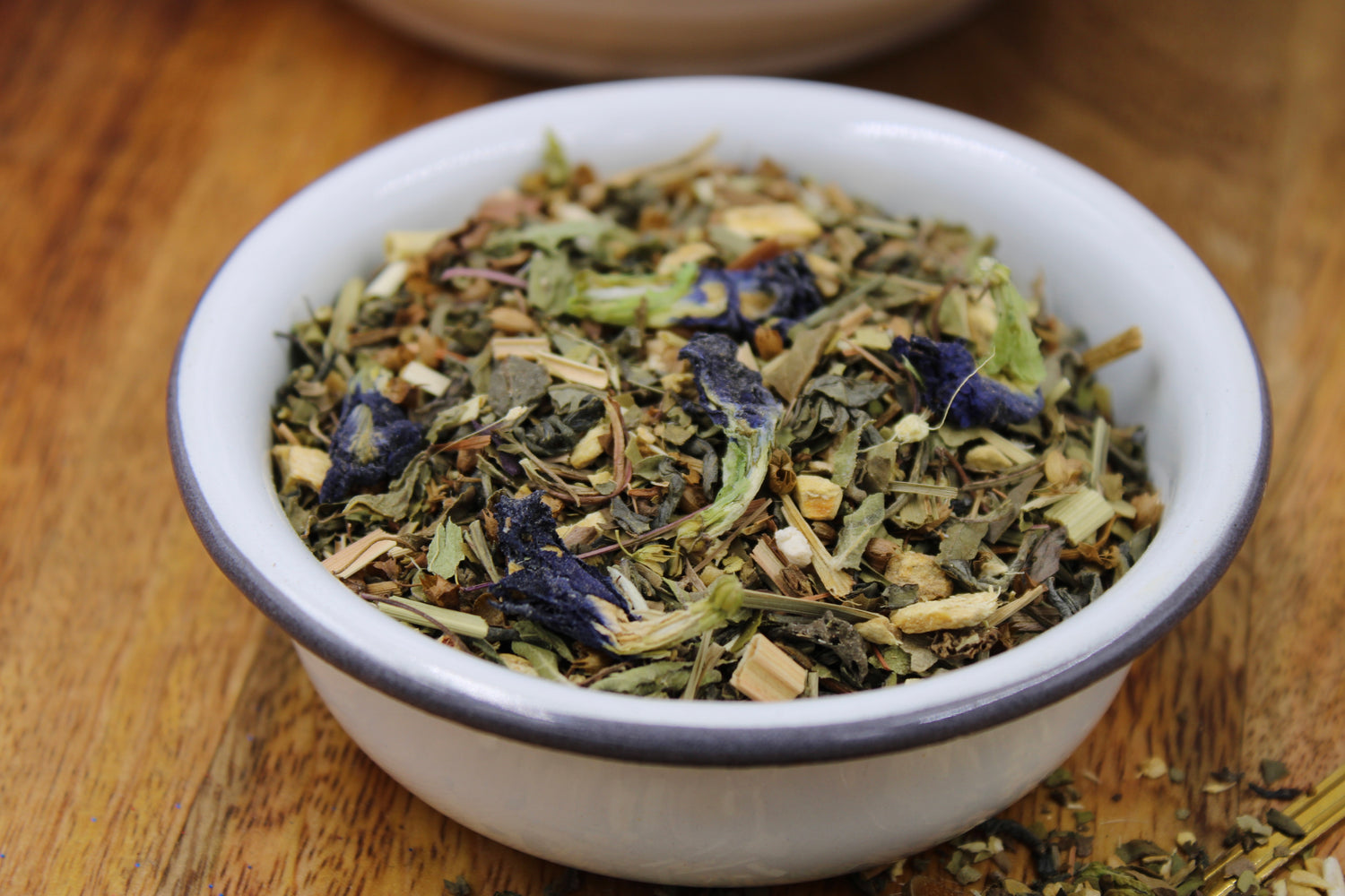 Good Vibes Tea blend by Relaxation Tea displayed in a white bowl on a wooden surface, showcasing its vibrant loose-leaf ingredients, including Moroccan mint, green tea, ginger, holy basil, lemongrass, yerba mate, butterfly pea flower, gotu kola, eleuthero, and echinacea. This refreshing and invigorating blend is crafted to uplift your mood and promote overall wellness, emphasizing Relaxation Tea’s commitment to high-quality, natural ingredients for health-conscious tea lovers.