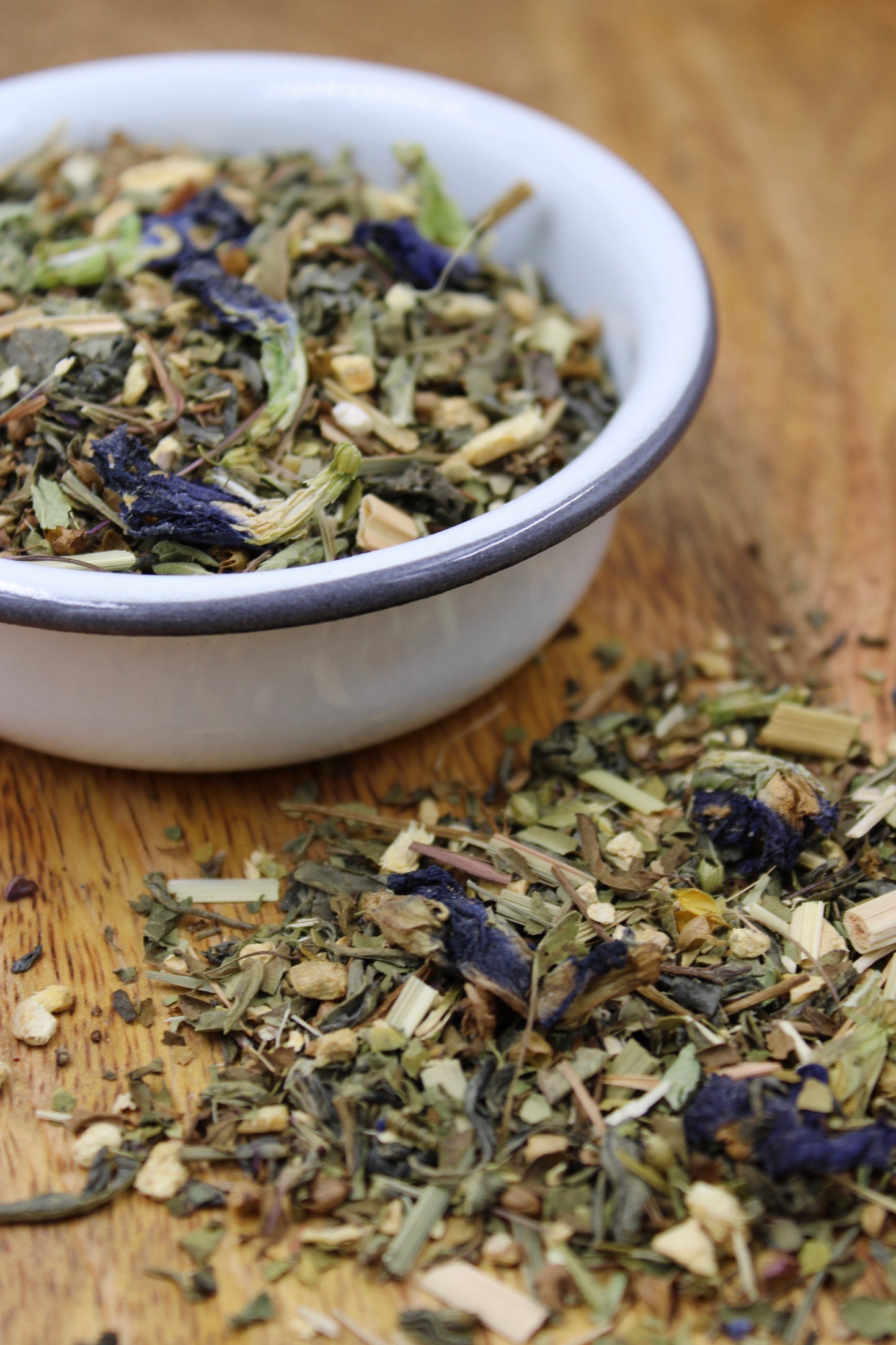 Organic Happy Vibes Tea blend by Relaxation Tea displayed in a white bowl with loose-leaf herbs spilling onto a wooden surface. Featuring organic Moroccan mint, green tea, ginger, holy basil, lemongrass, yerba mate, butterfly pea flower, gotu kola, eleuthero, and echinacea, this vibrant blend is designed to uplift your mood and promote overall wellness, reflecting Relaxation Tea’s dedication to high-quality, organic ingredients and holistic health.